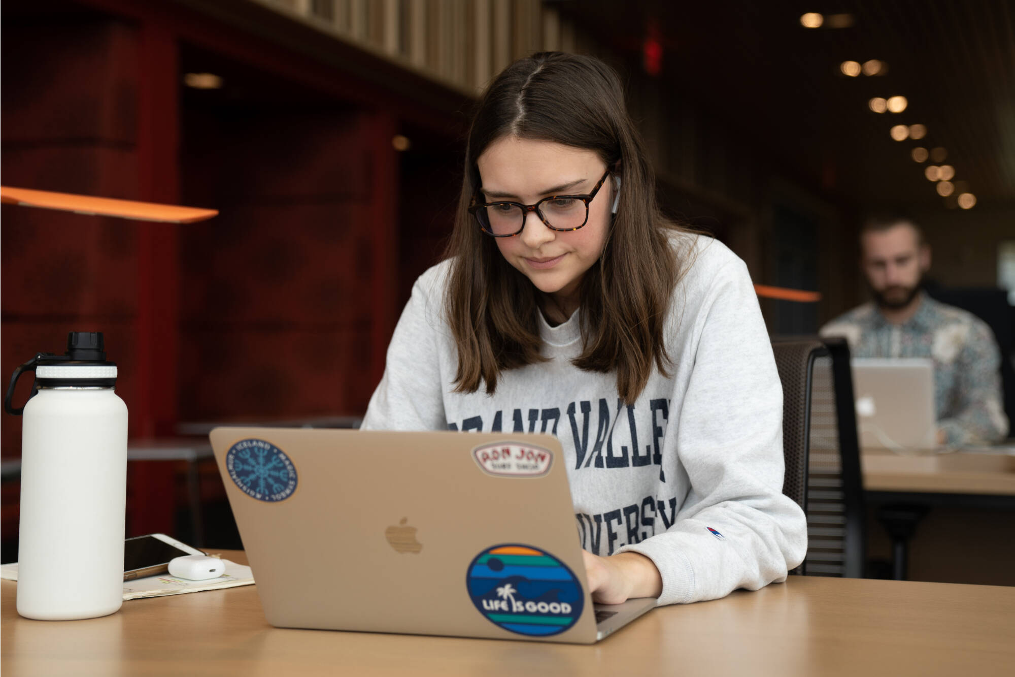 Grand Valley student sitting on a desk at the Pew Library working on their laptop. They are wearing a Grand Valley sweatshirt.